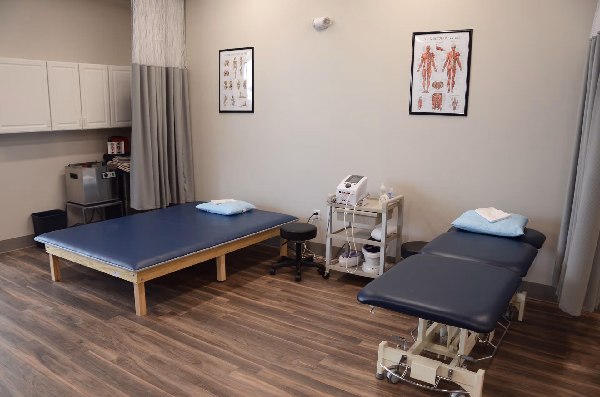 Two treatment tables in a calm physical therapy room with equipment and posters.