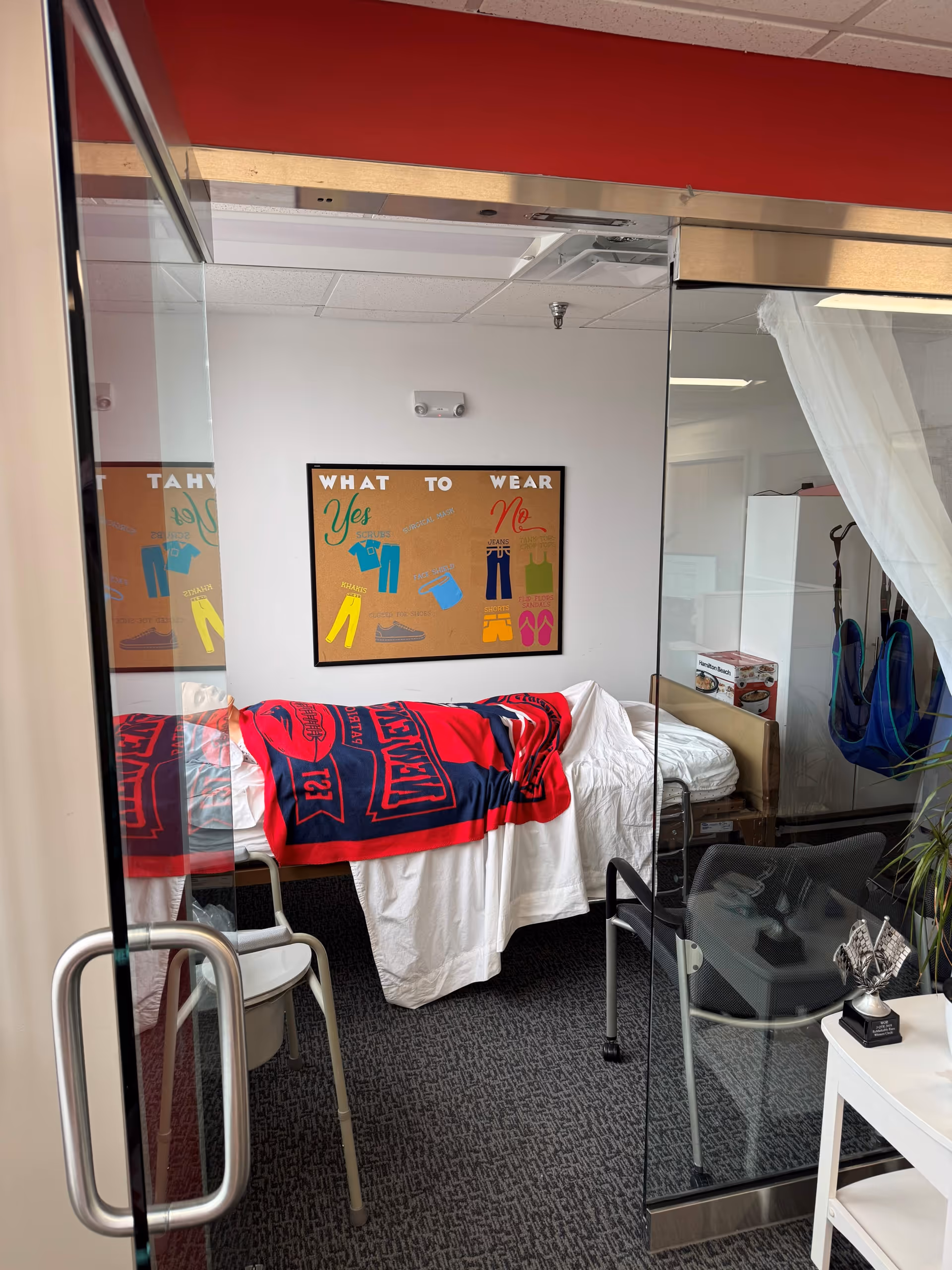 Hospital-style bedroom with bed, red blanket, posters, and glass doorway in foreground.