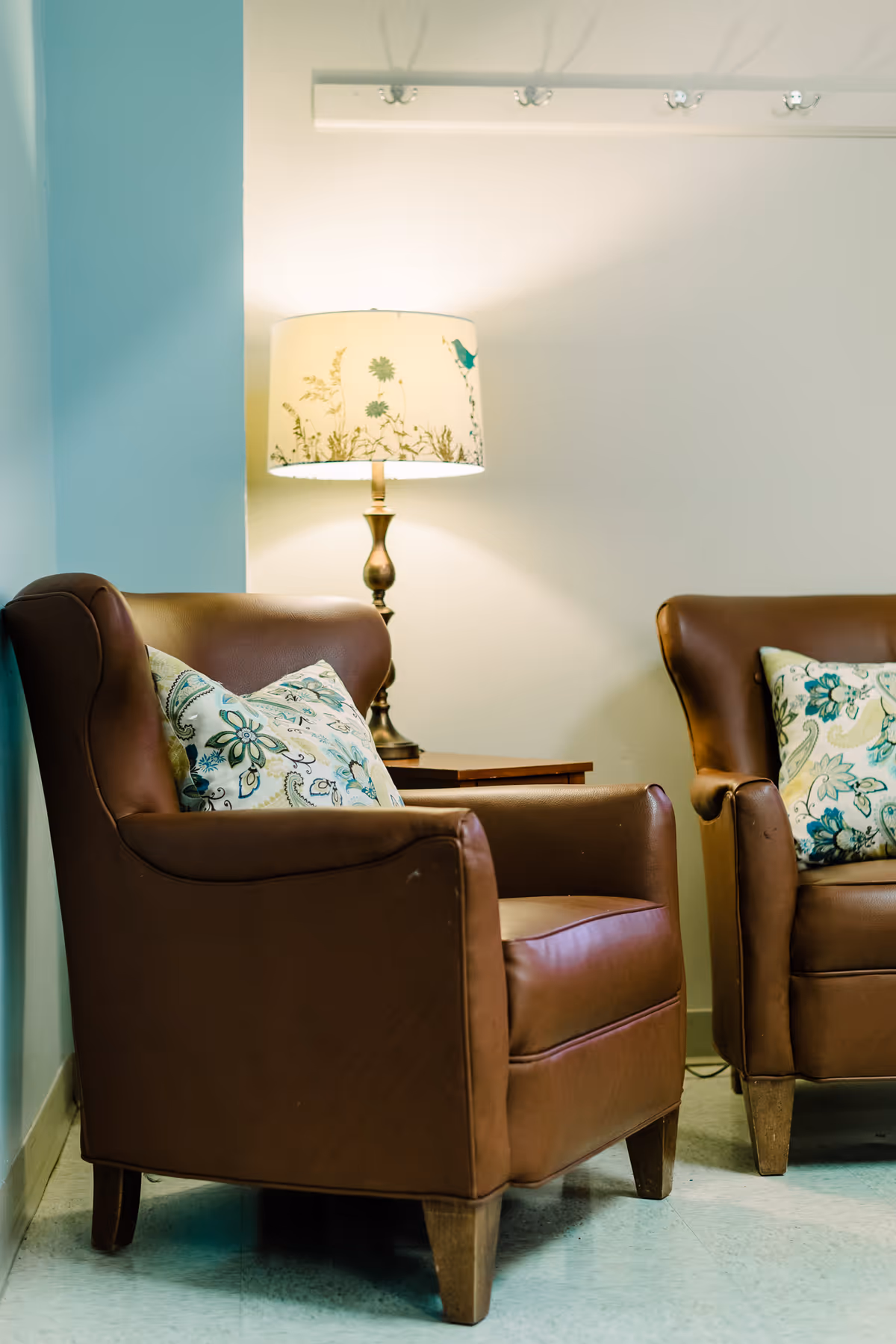 Two brown leather armchairs with patterned pillows sit beside a lamp in a pastel living area.