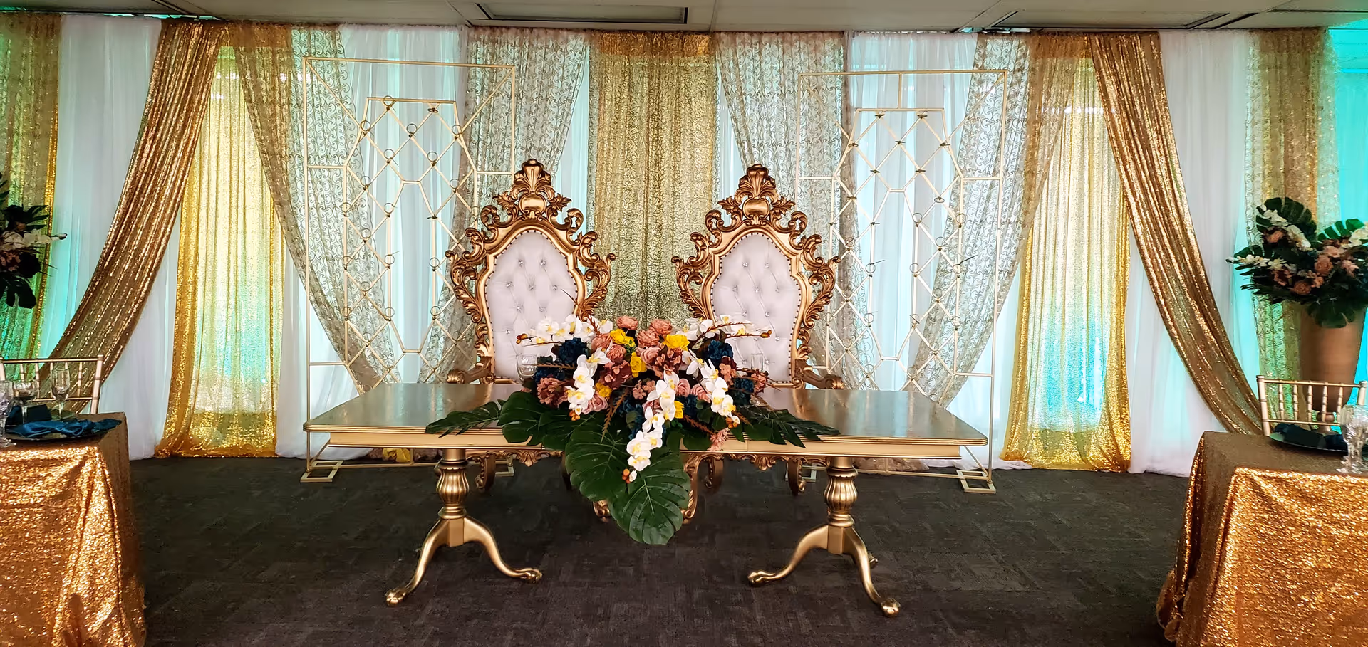 Elegant indoor stage setup with ornate gold chairs, floral centerpiece, and shimmering drapes.