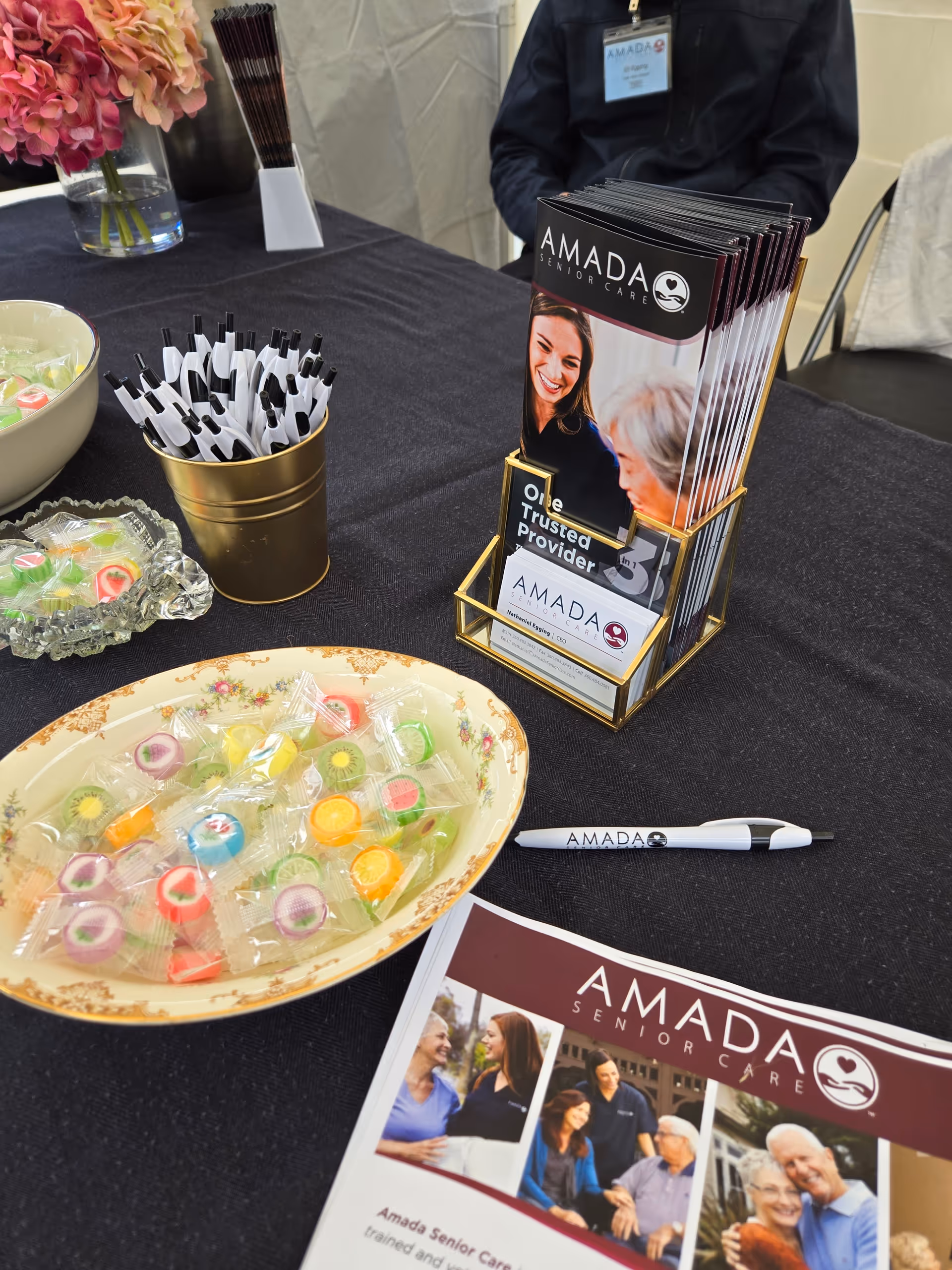 Tabletop display at Amada Senior Care booth with brochures, pens, candy, and a staff member.