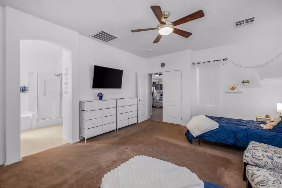 White-walled bedroom with blue bedding, dresser, TV, and adjacent bathroom.