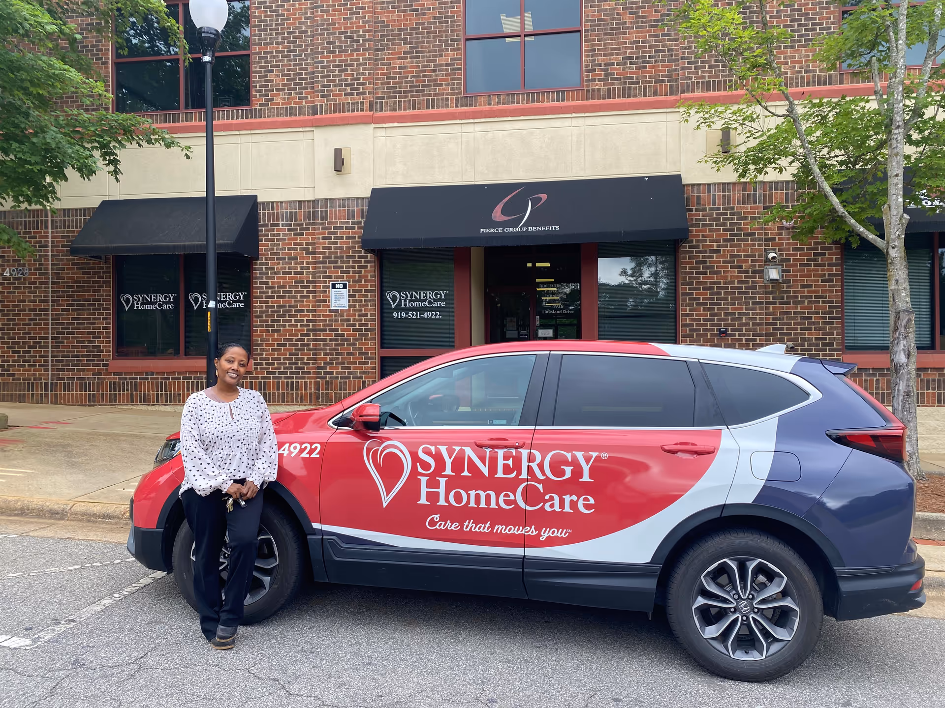 A person stands beside a red Synergy HomeCare van parked in front of brick facility.