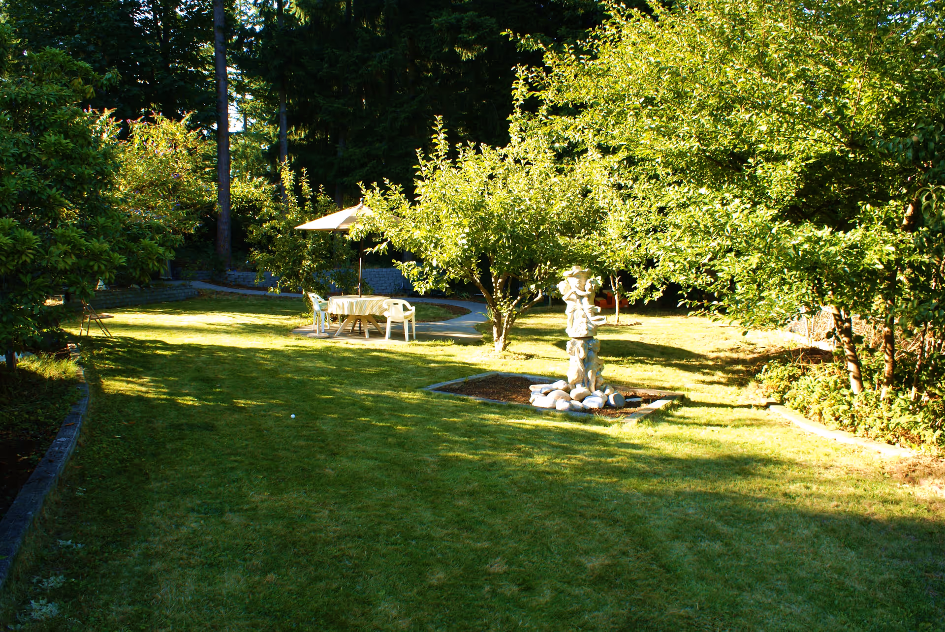 A sunny grassy courtyard with a table under an umbrella, chairs, and a stone statue fountain.