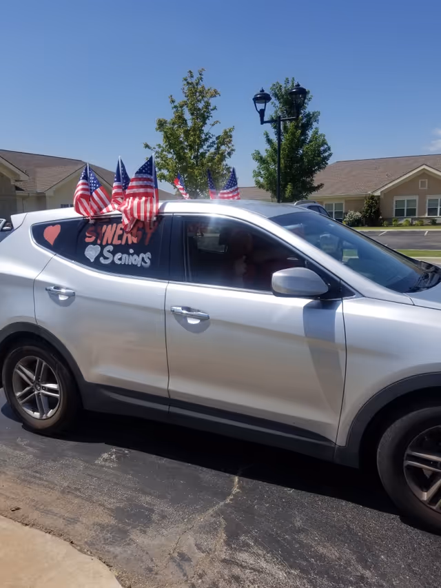Silver SUV branded with SYNERGY HomeCare and patriotic flags in a parking lot.