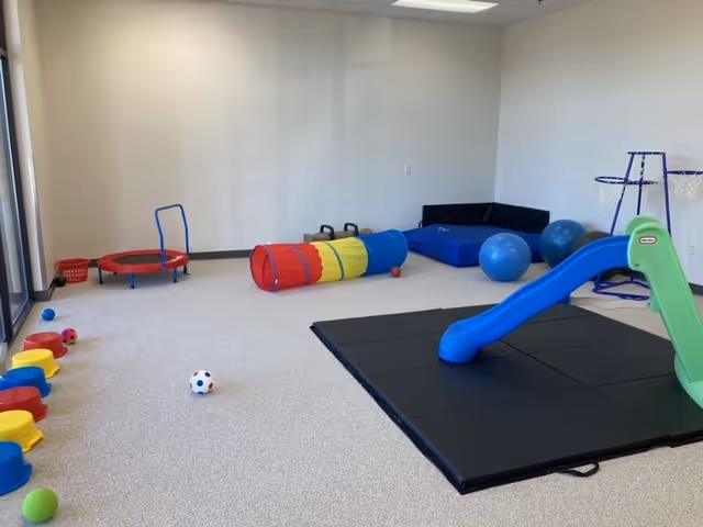 Indoor playroom with a blue slide, soft mats, tunnel, balls, and colorful play equipment.