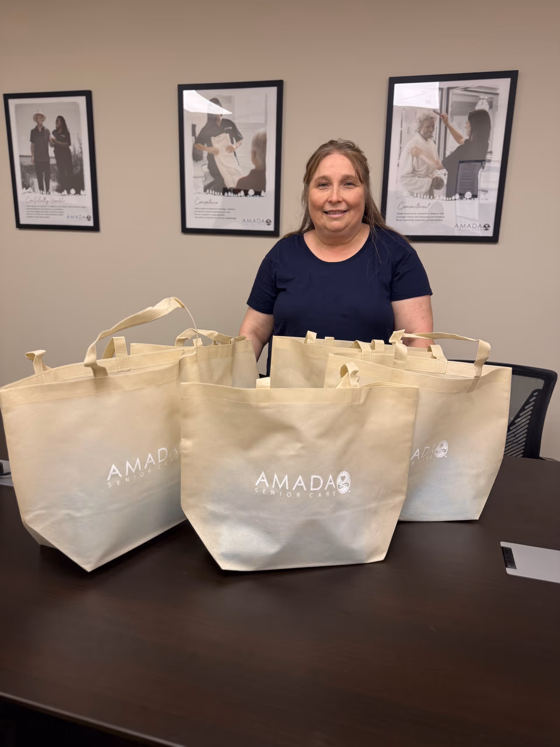 Smiling staff member stands behind a table with beige Amada tote bags in a conference room.