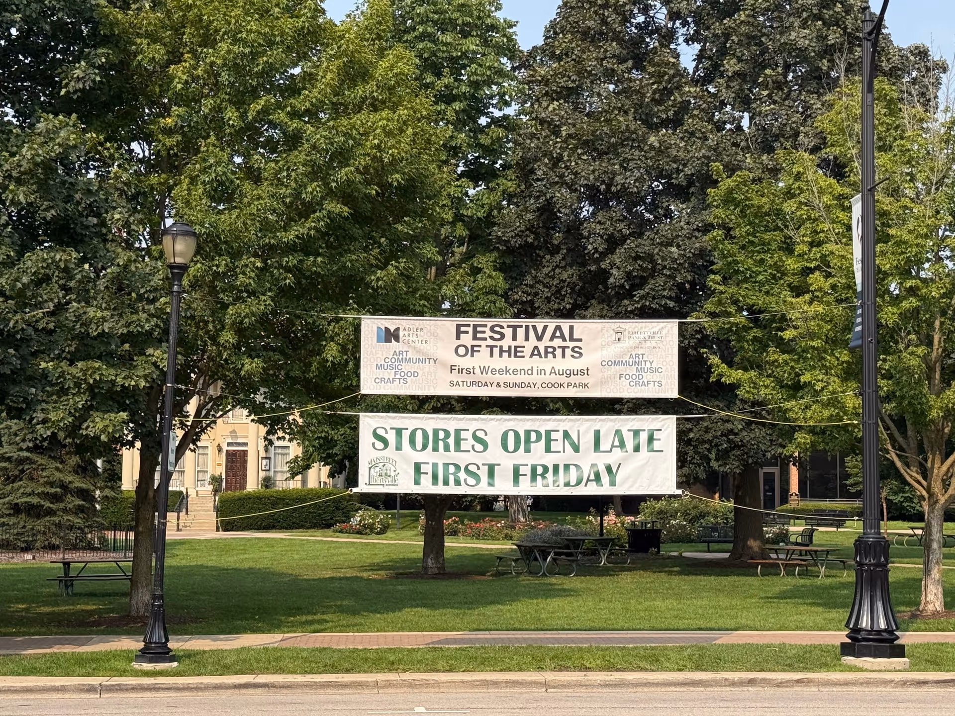 Outdoor park area with banners advertising arts festival, trees, benches, and lampposts.