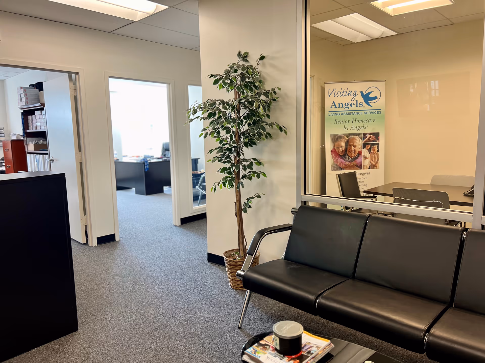 Office lobby with black leather sofa, potted tree, glass partition, and Visiting Angels banner.