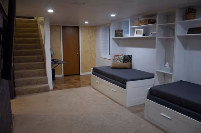 Basement-like bedroom with built-in beds, shelving, and a small desk under stairs.
