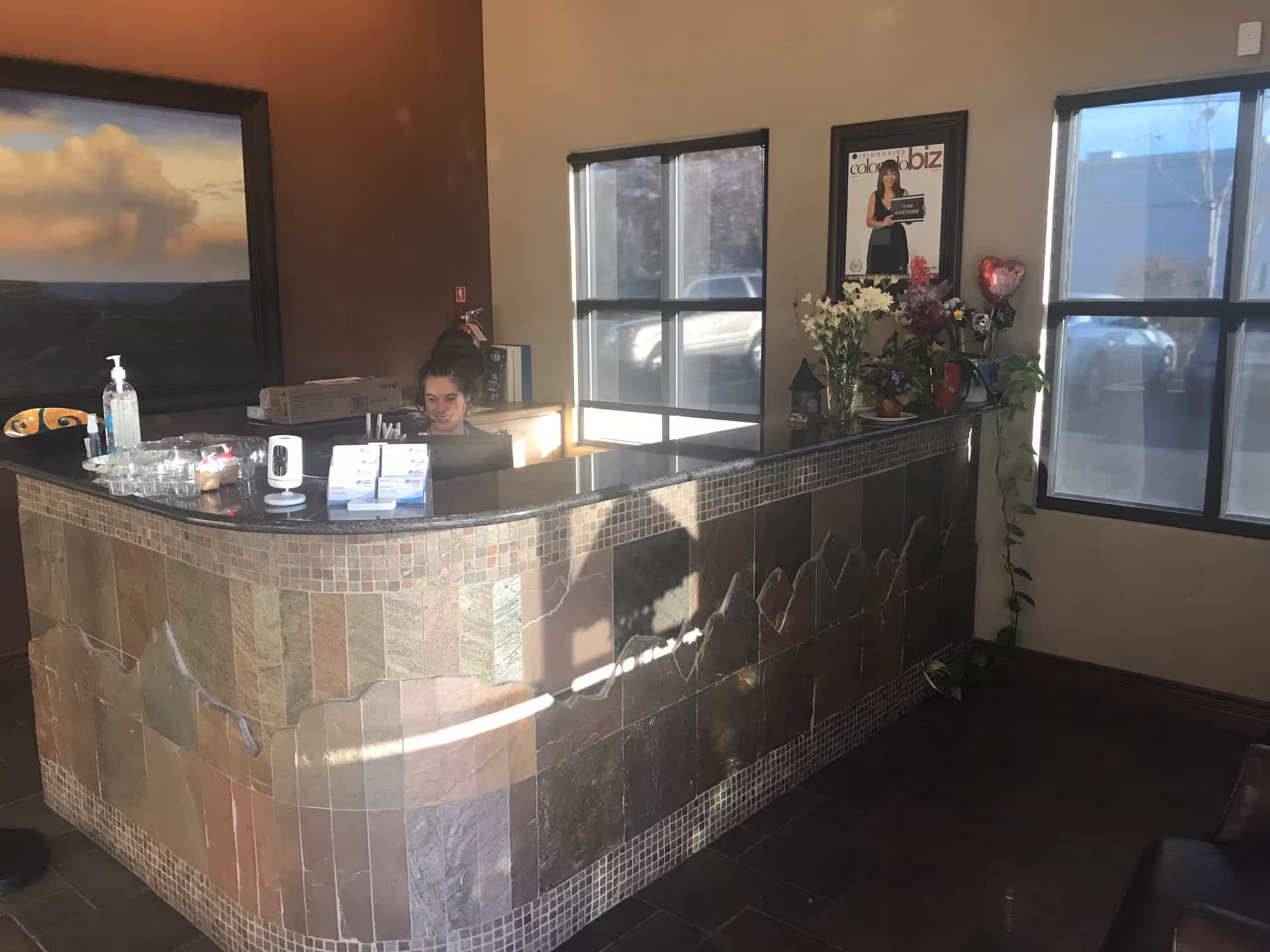 Reception desk with a staff member, tiled front, flowers, and windows in lobby.
