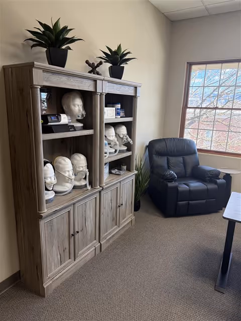 Wooden cabinet with mannequin heads, a black recliner, plants, and a window.
