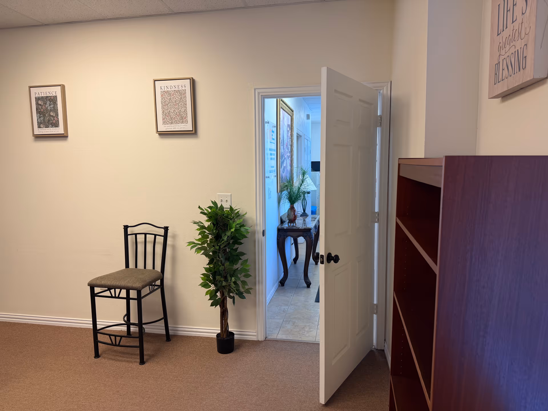 A neutral hallway with a chair, potted plant, and open door to a back room.