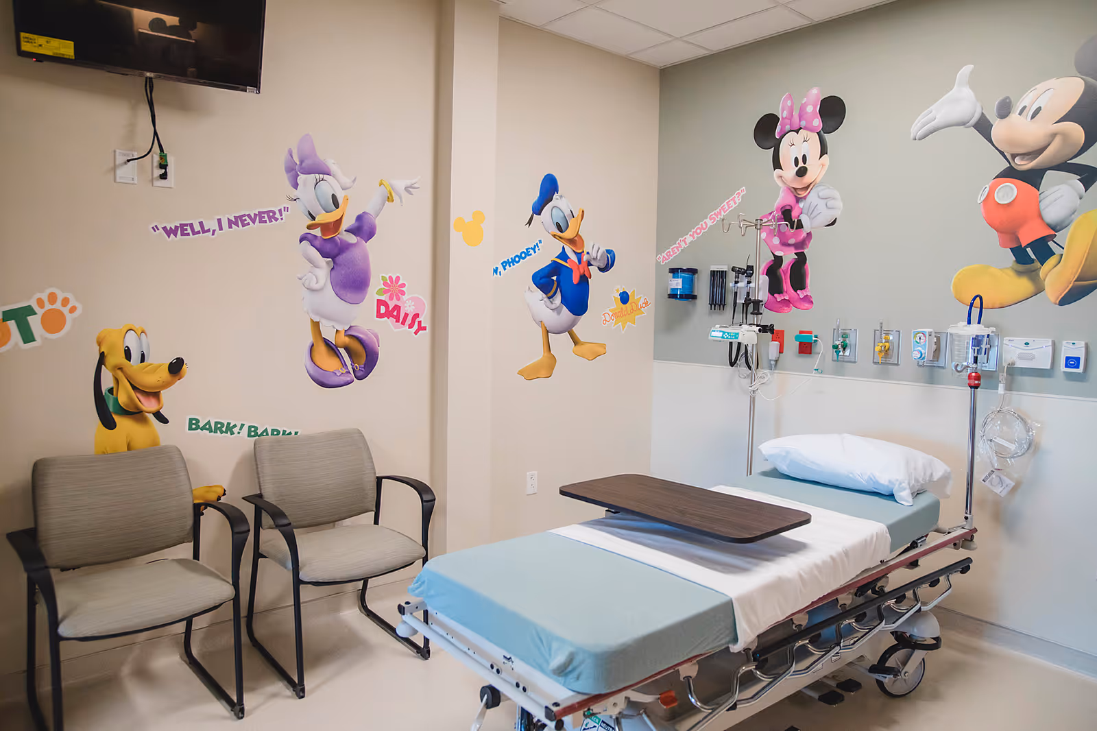 Pediatric hospital room with cartoon character wall decals, a medical bed, and seating along the wall.