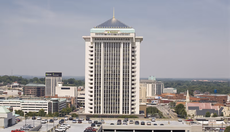 Tall modern high-rise building with surrounding cityscape and a parking lot in foreground.