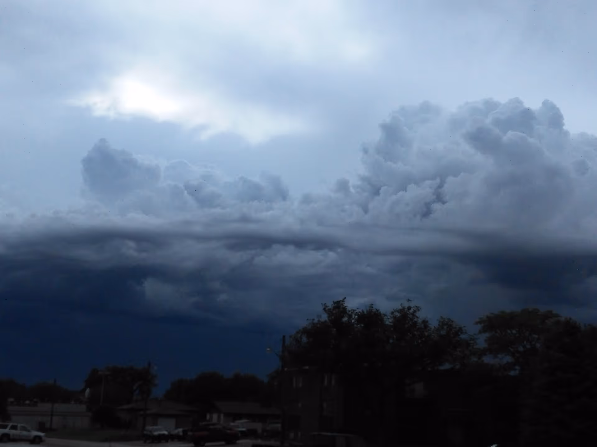 Dark storm clouds loom over a suburban neighborhood with silhouetted trees and buildings.