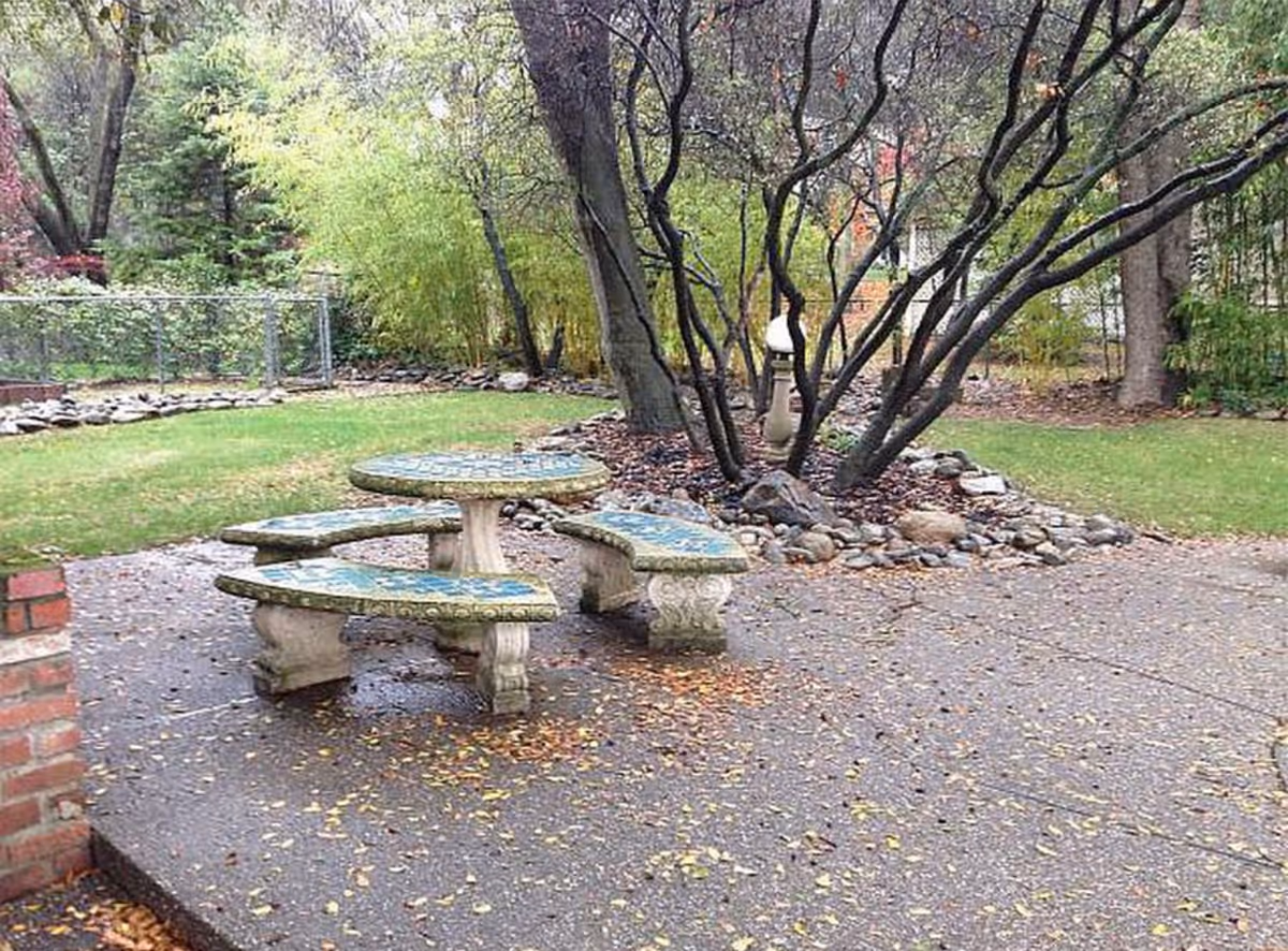 Outdoor garden area with a stone round table and benches, surrounded by trees and greenery.