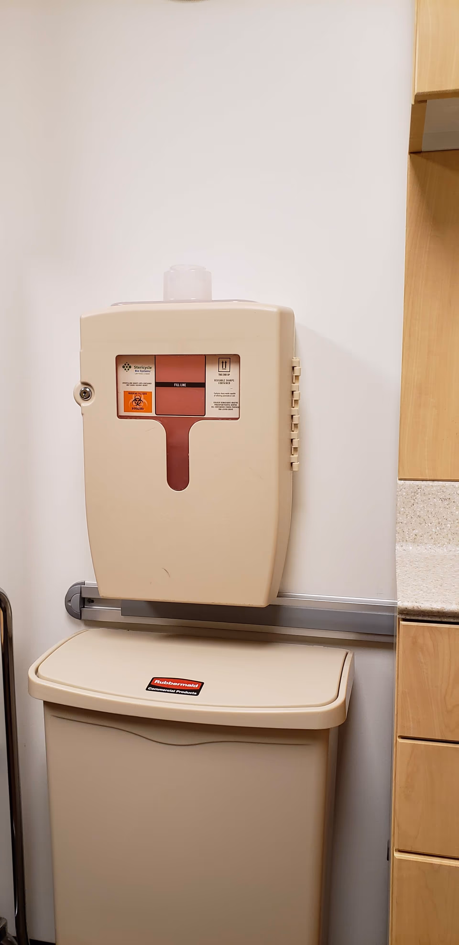 Wall-mounted medical waste/sharps container above a closed beige disposal bin in a clinical kitchen area.