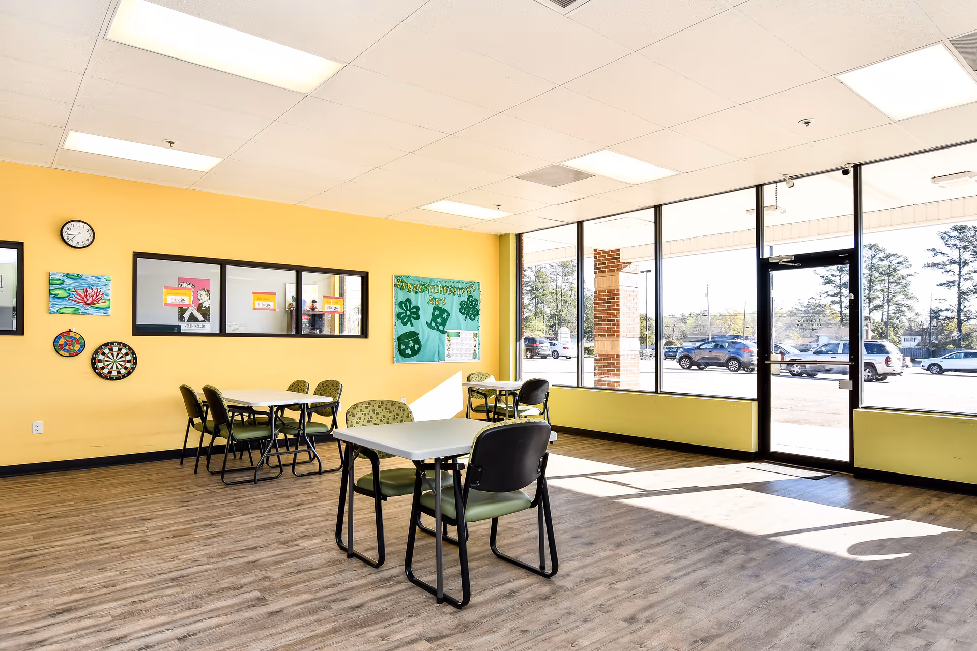 Bright interior community room with yellow walls, large windows, tables, chairs, and colorful wall art.