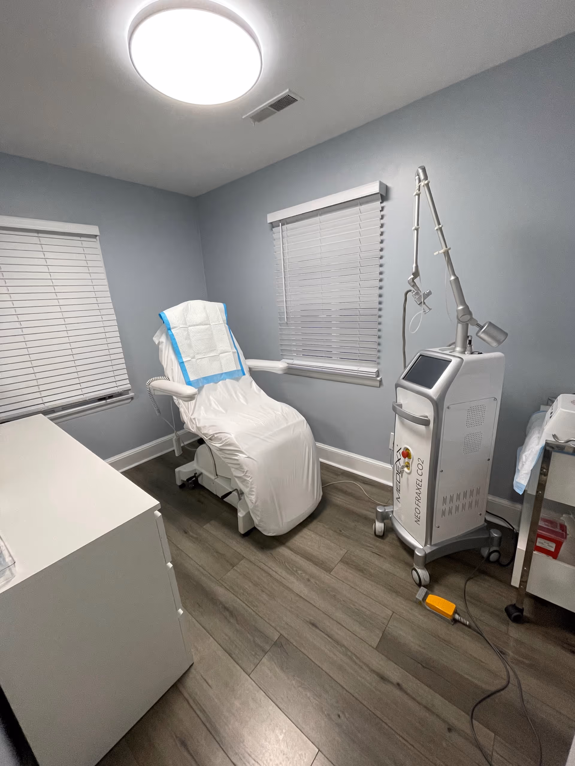 Treatment room featuring a covered clinic chair, a medical device on wheels, and window blinds.