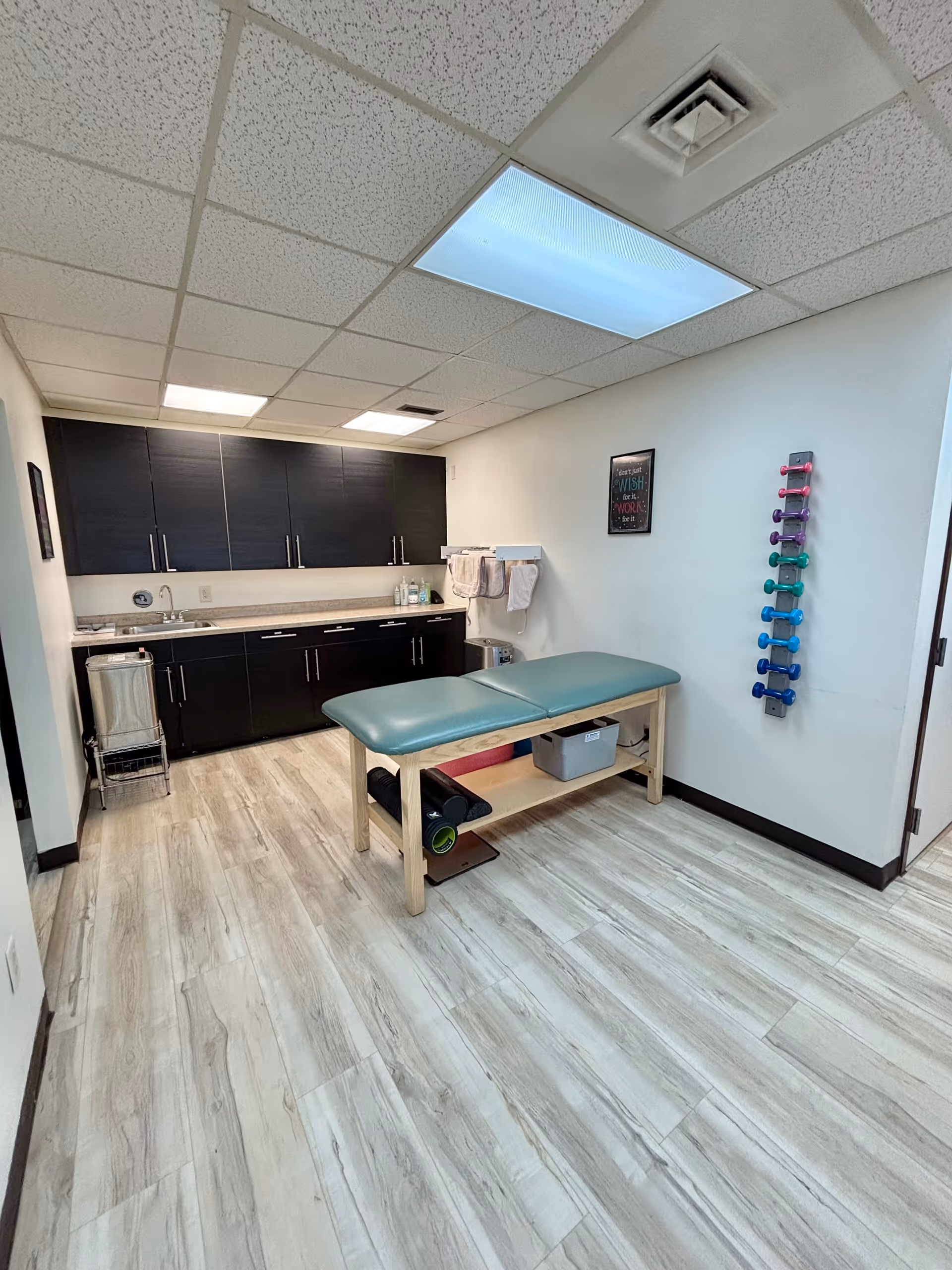 Therapy room with teal examination table, dark cabinets, sink, and exercise equipment.