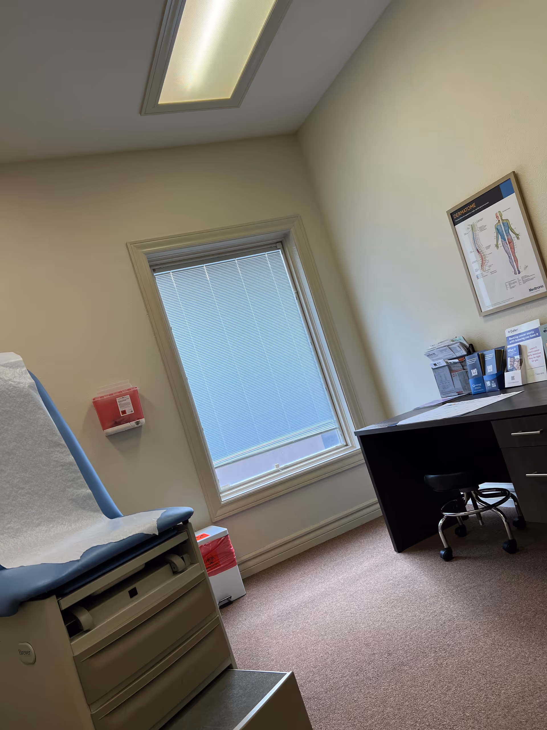 Medical examination room with exam chair, window, and desk.