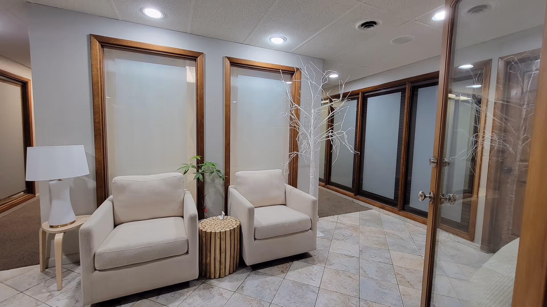 A modern lobby area with two beige armchairs, a lamp, and glass doors.