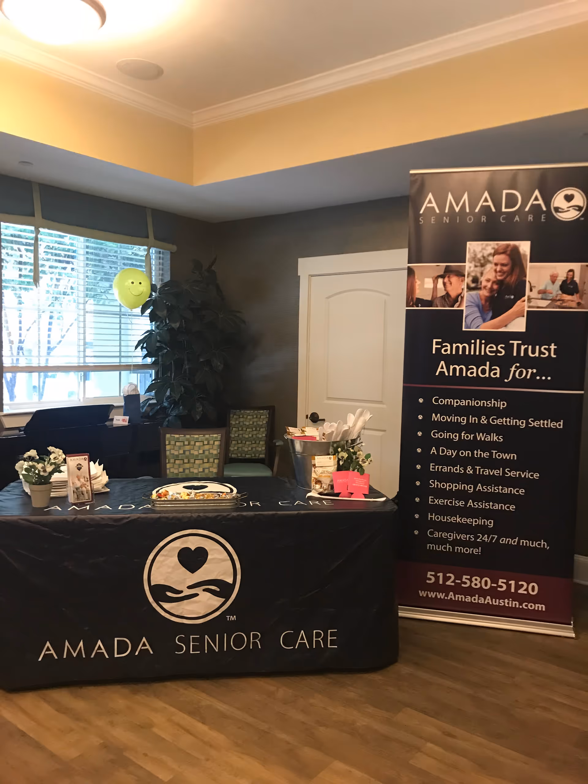 Indoor event setup with Amada Senior Care banner, table, cake, balloons, and plants.