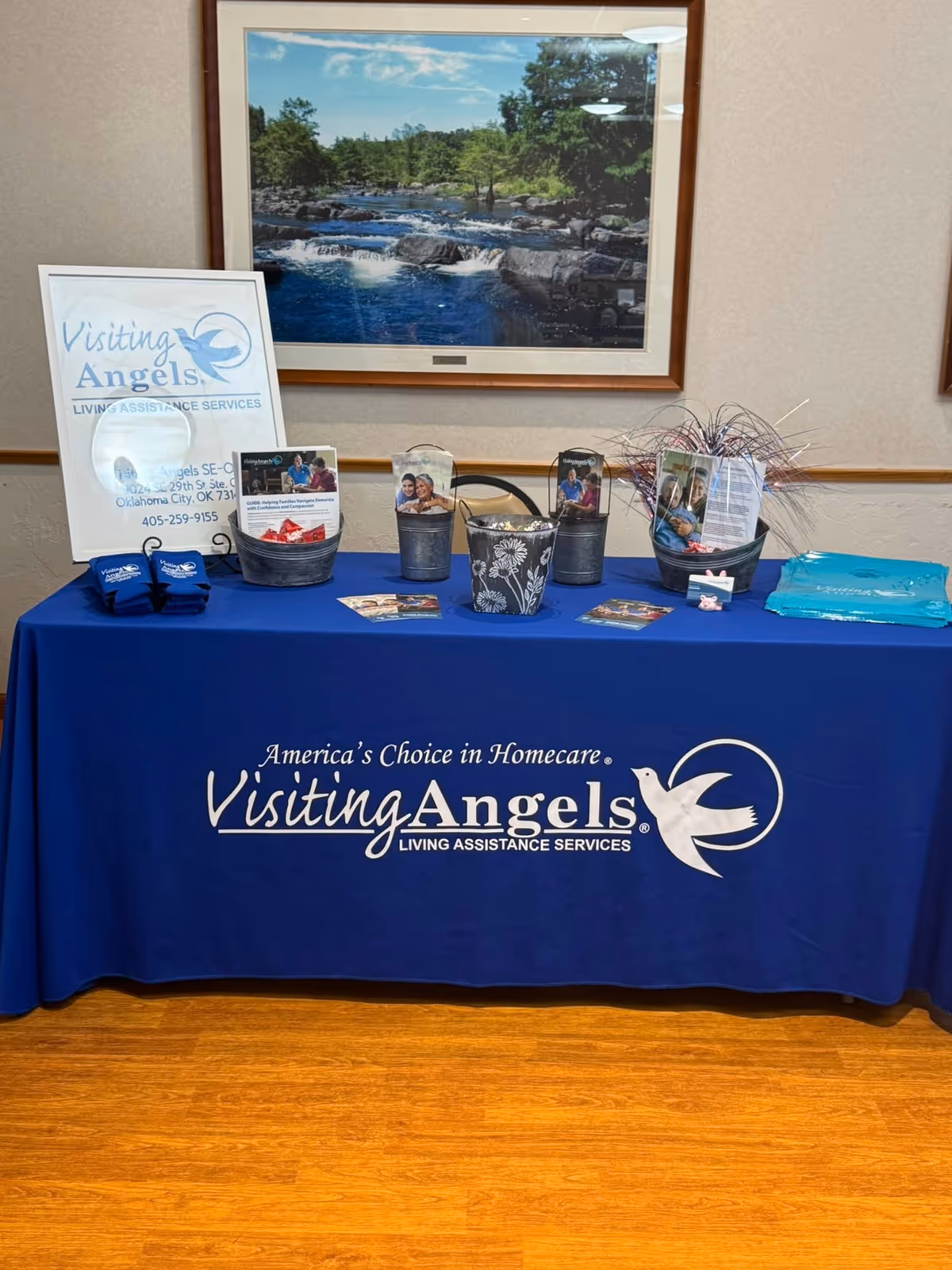 Indoor display table with blue Visiting Angels cloth, brochures, buckets, and a framed wall photo.