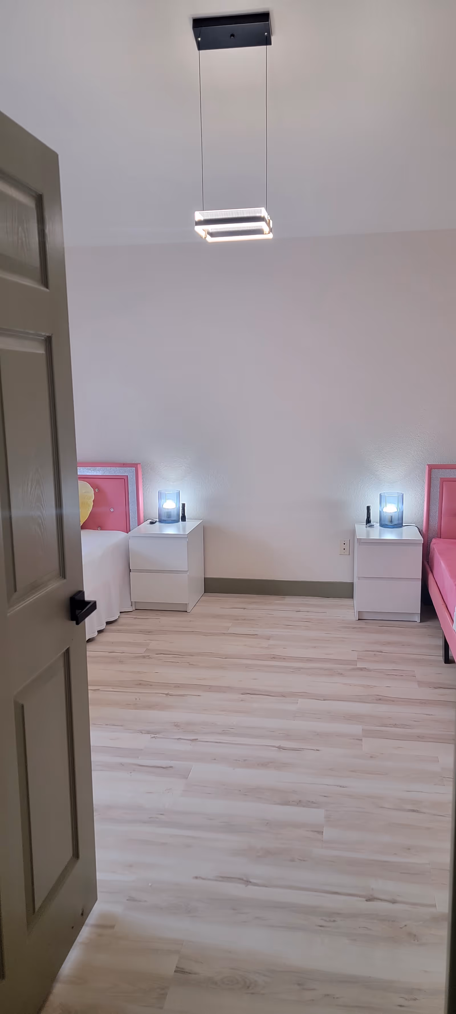 Two twin beds with pink headboards, white nightstands, and glowing lamps in a bright bedroom.