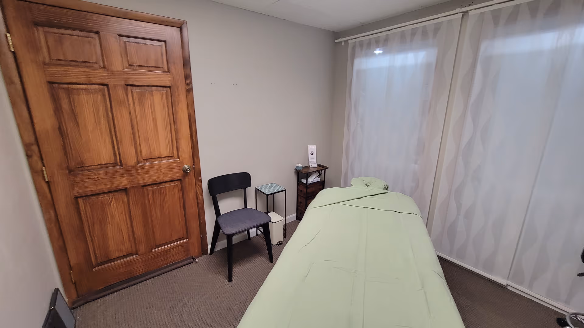 A massage treatment room with a green-covered table, chair, small side table, and sheer curtains.