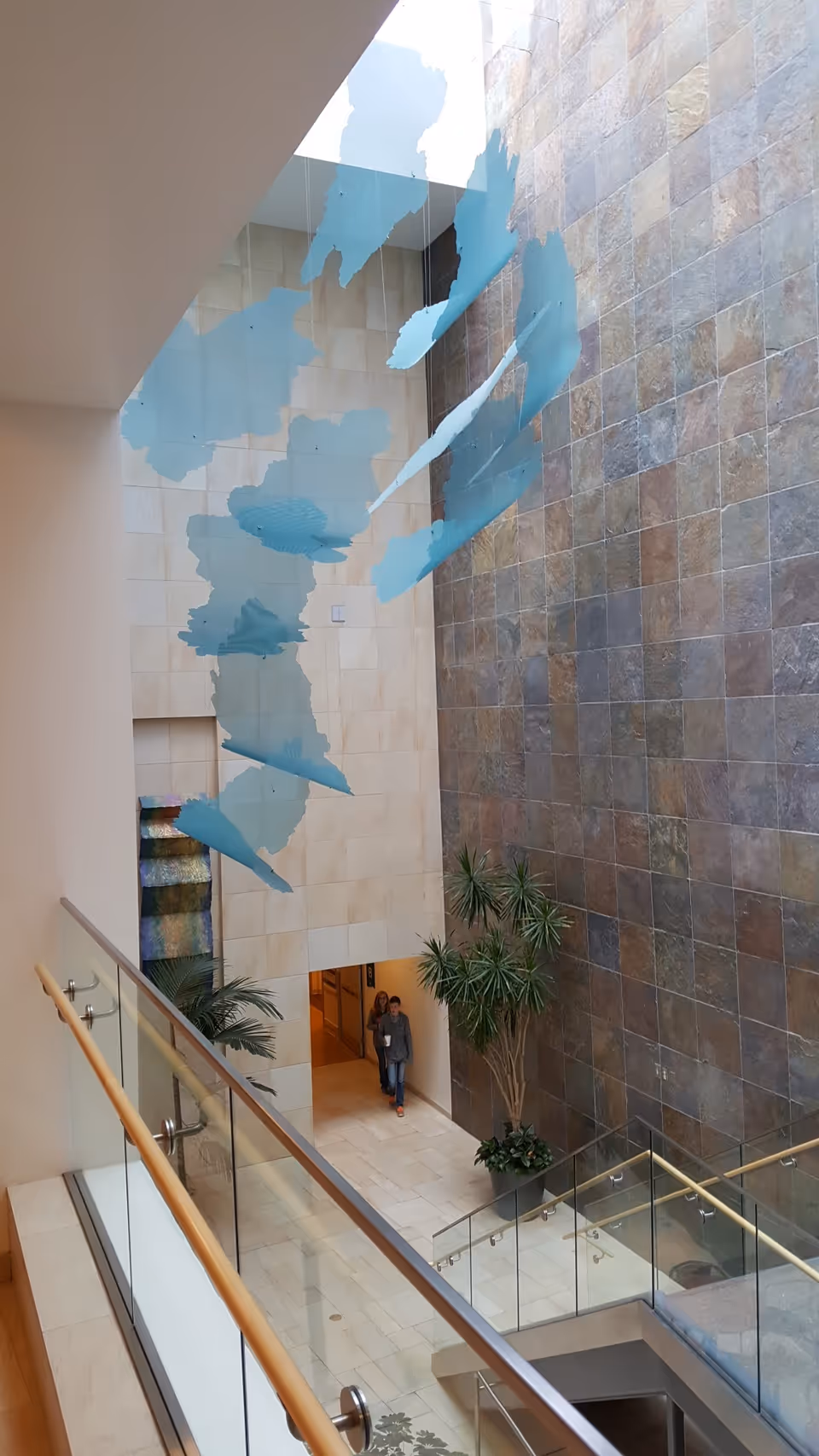 Interior atrium with glass railing, stone wall, plants, and two people entering.