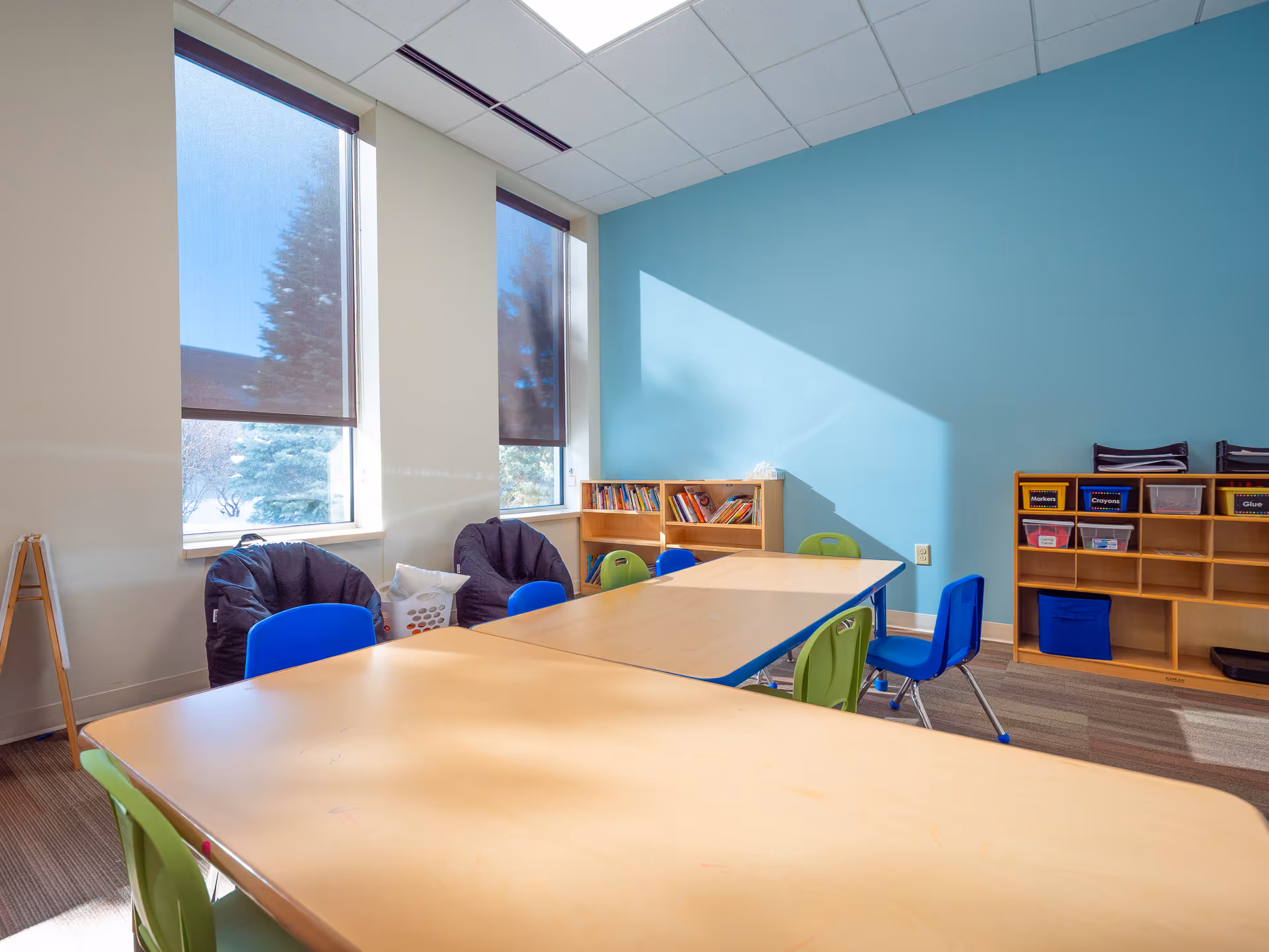 Bright classroom-style room with tables, colorful chairs, and shelves along teal wall.