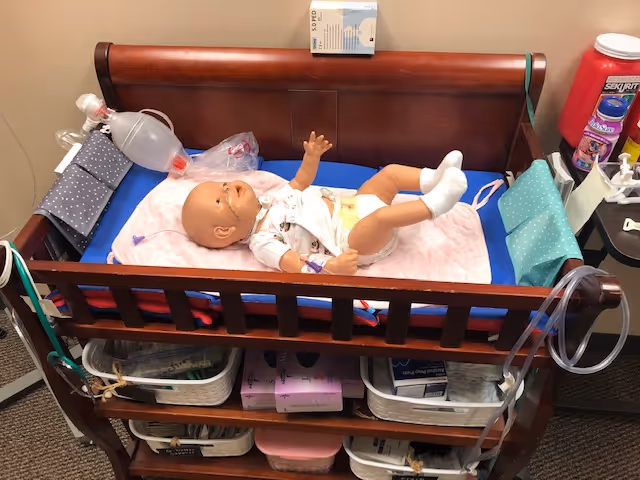 A wooden infant crib with a plastic baby doll and medical supplies in a clinical room.