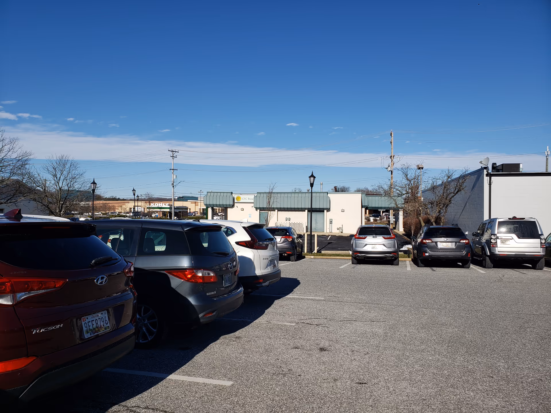 Parking lot full of cars with a low commercial building and blue sky in background.