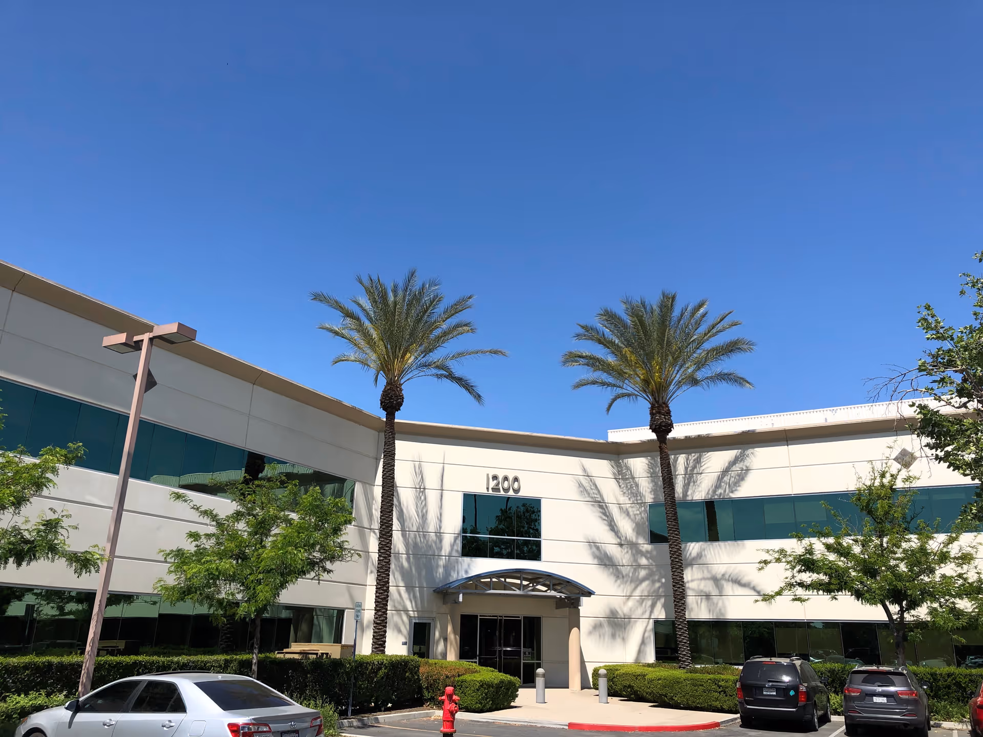 Front exterior of a modern building with palm trees and parked cars under a blue sky.