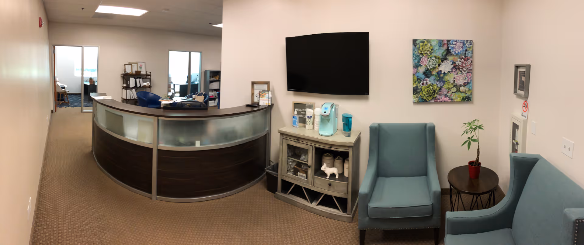 Curved reception desk in a lobby with teal chairs, a TV, and decorative plant.