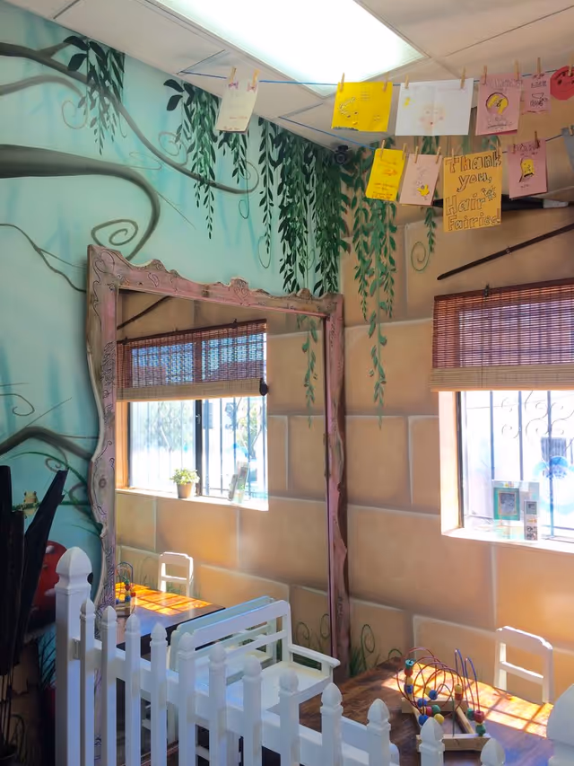 Colorful whimsical indoor playroom with vine mural, pink ornate mirror, white picket fence, and toys.