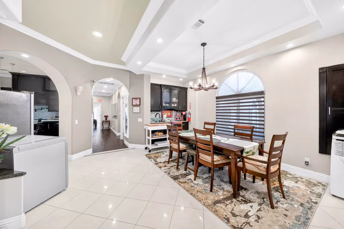 A bright dining area with a long wooden table, chairs, and archways in a senior home.