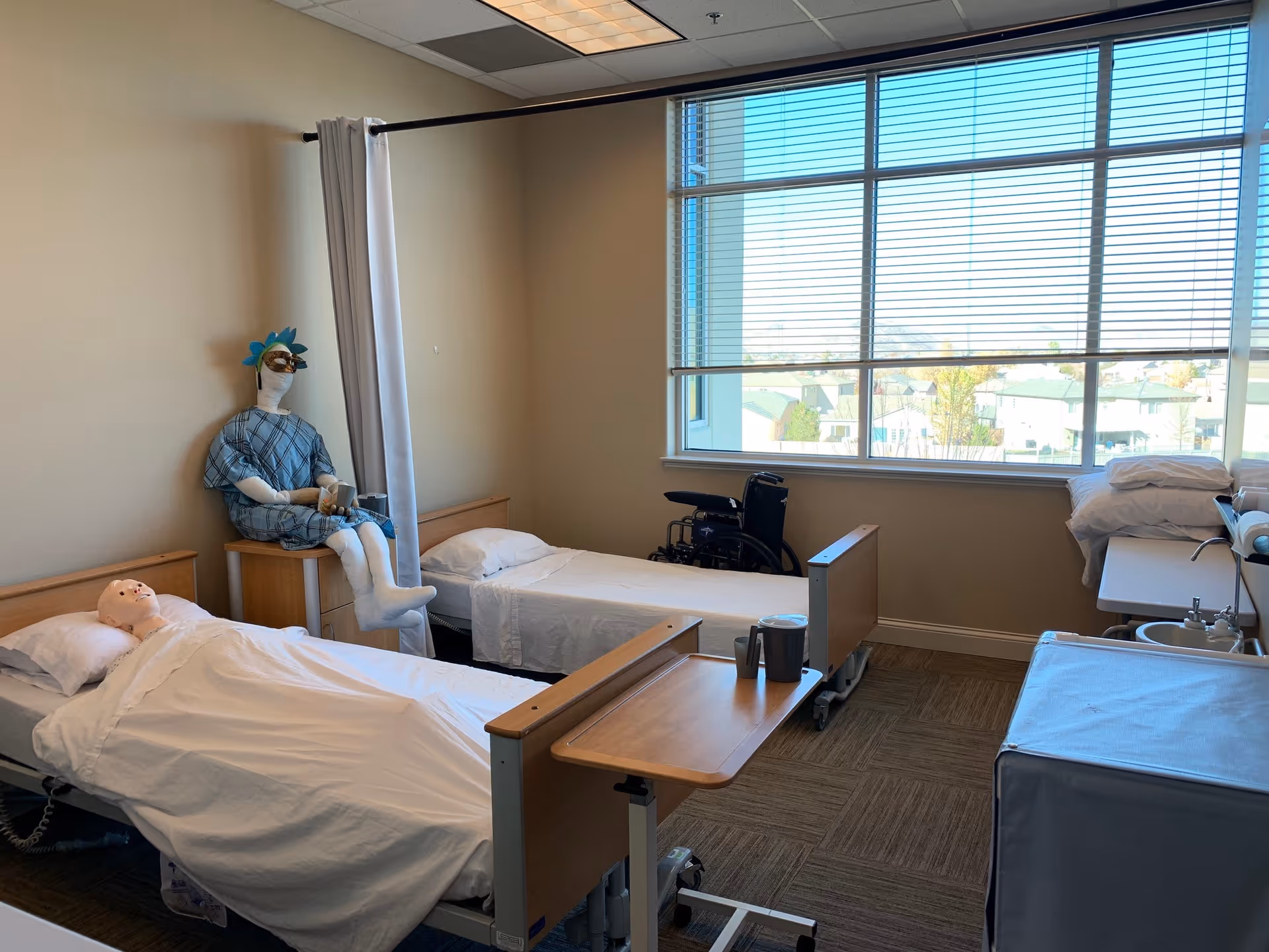 Bright patient room with two beds, a mannequin, wheelchair, and a large window.