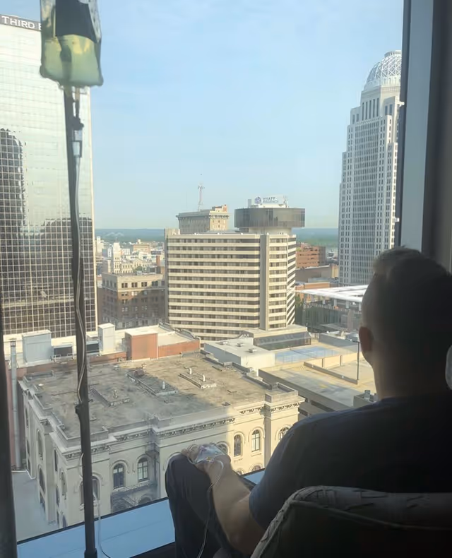 Resident sits by a large window overlooking a city skyline, with an IV line.
