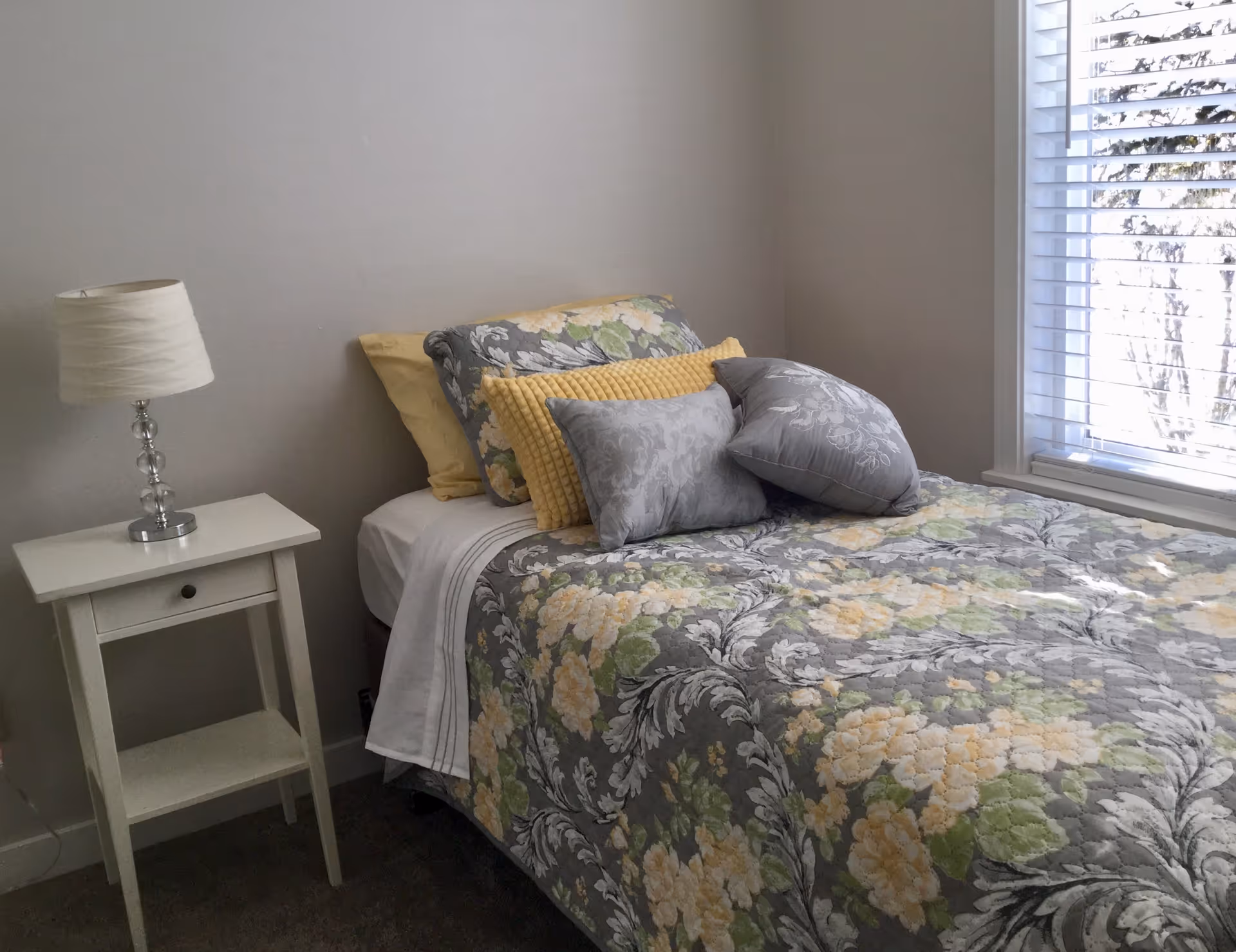 A neatly made bedroom with a floral bedspread, pillows, lamp, and window blinds.