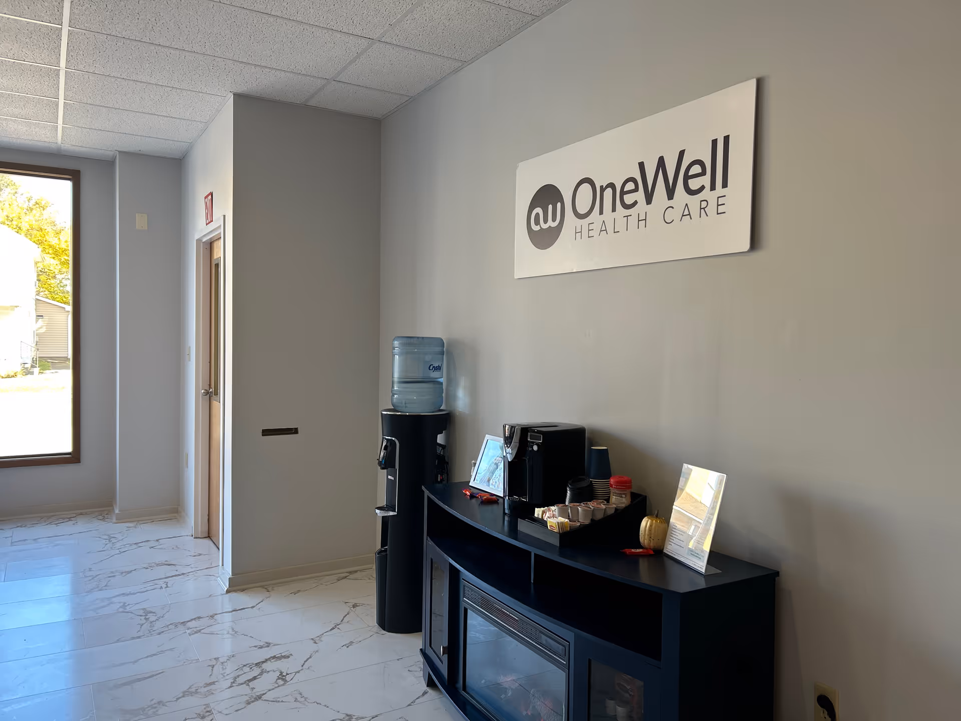 Facility lobby area with OneWell Health Care sign, water cooler, coffee station, marble floor.