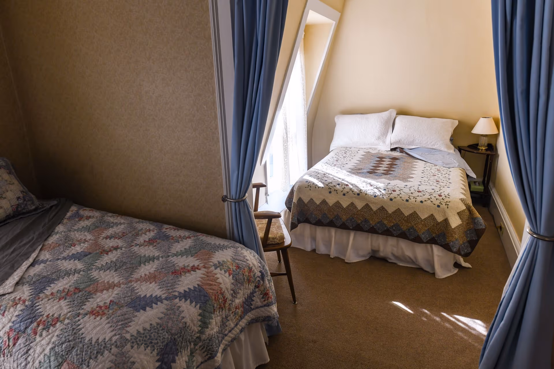 Two twin beds with quilts and blue curtains in a shared senior living bedroom.