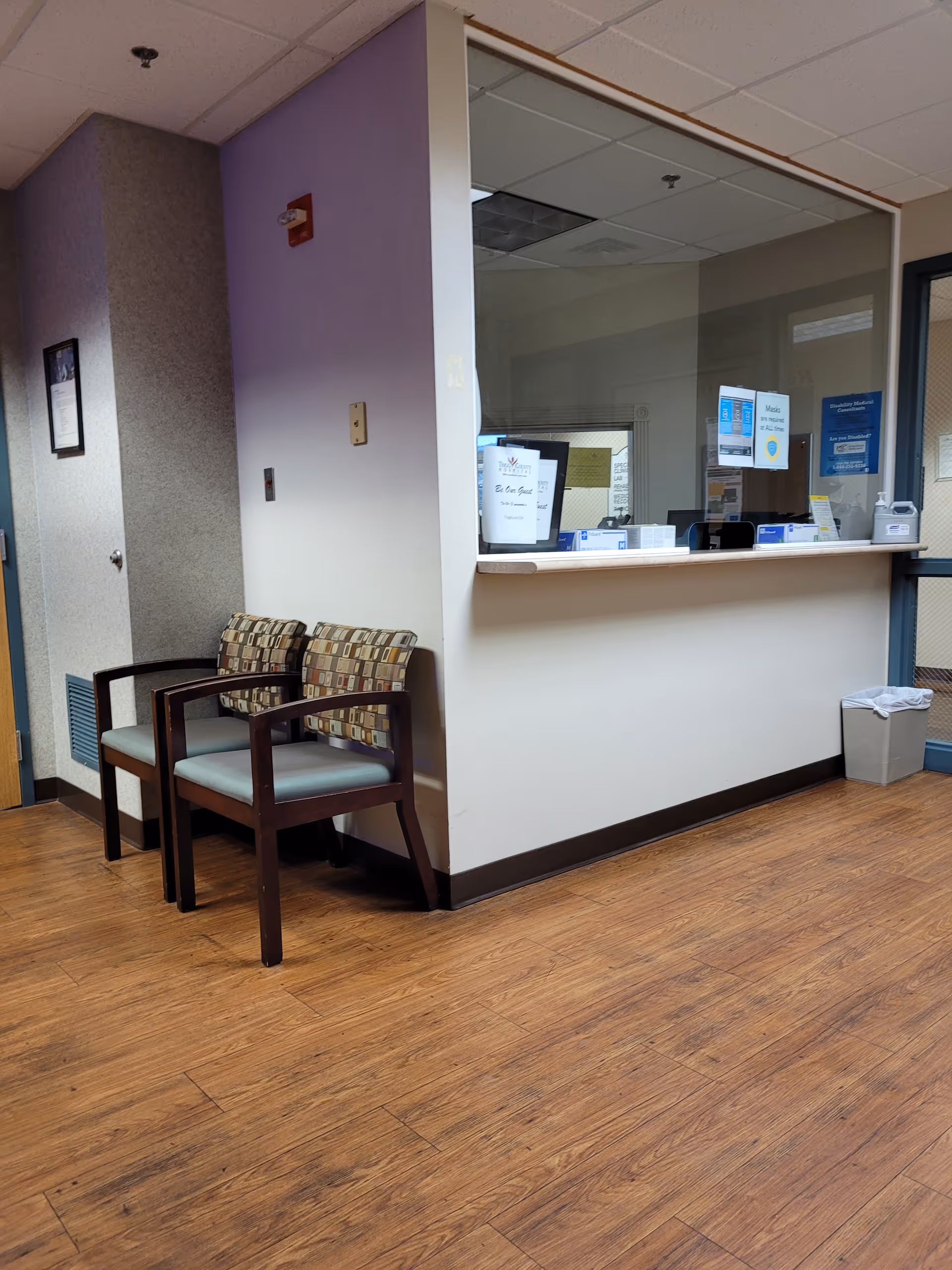 Waiting area with two patterned chairs beside a front desk window in a hospital lobby.
