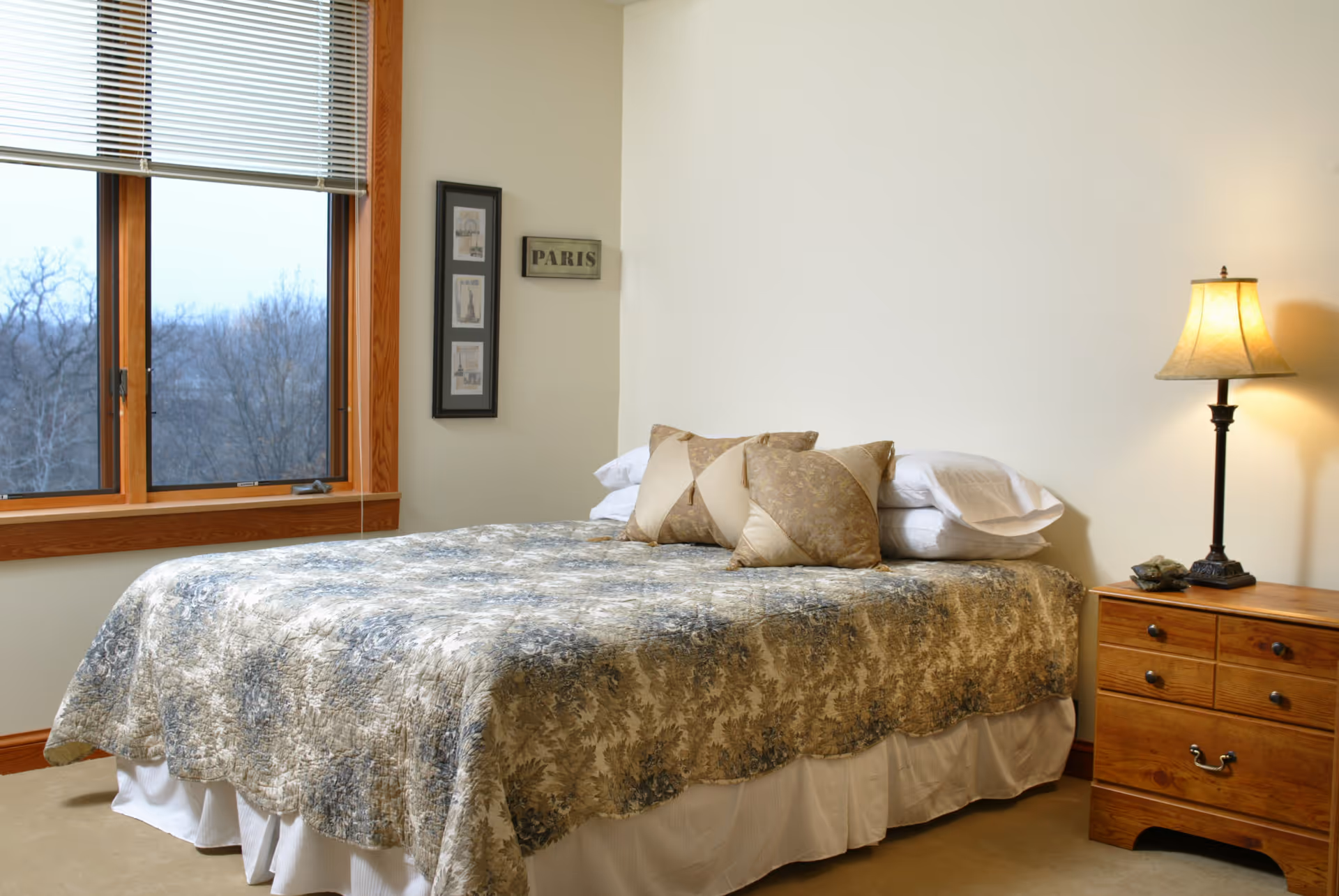 Cozy bedroom featuring a bed with a patterned quilt, pillows, nightstand, and a lit lamp.