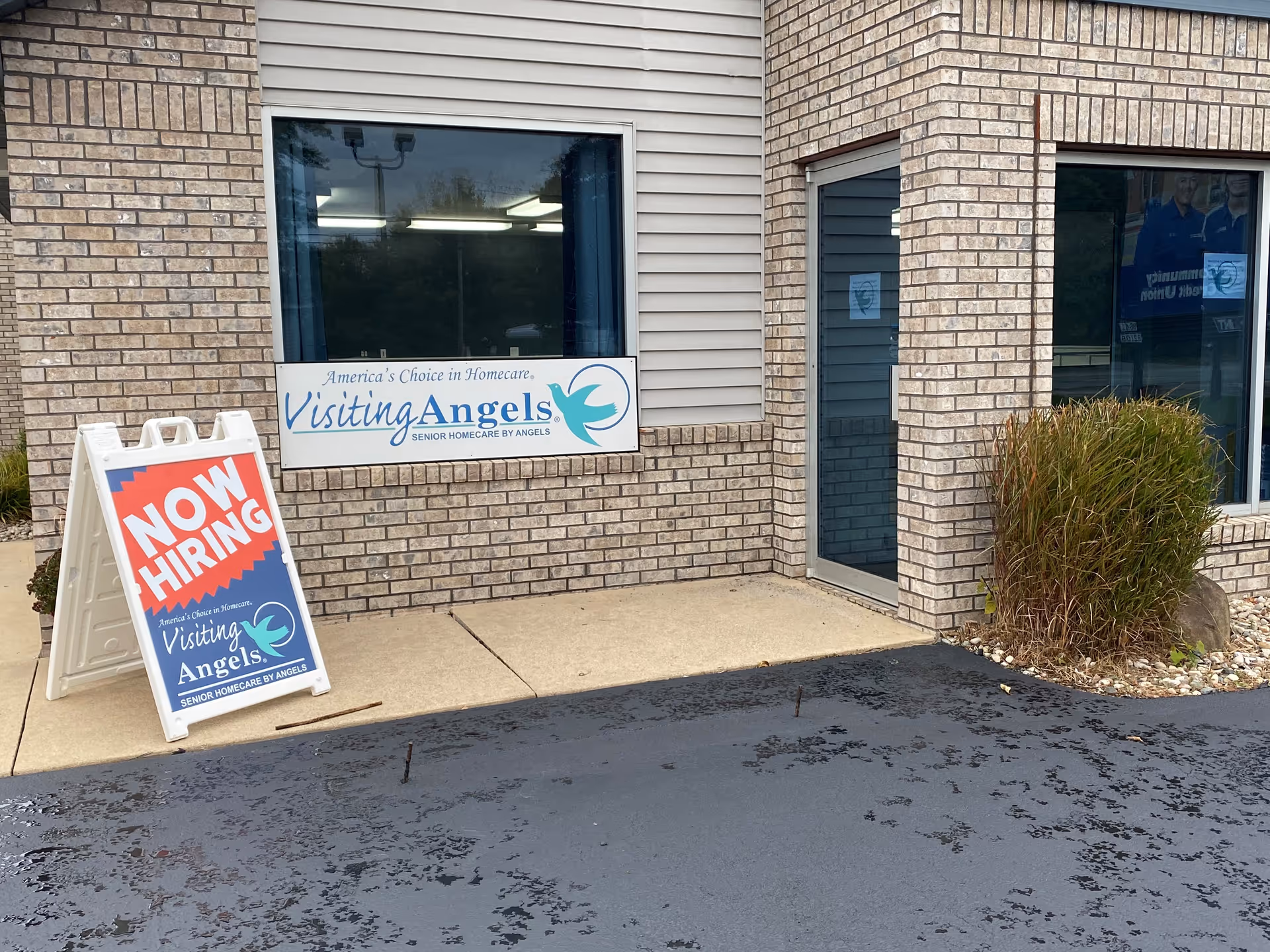Exterior brick building entrance with Visiting Angels sign and Now Hiring board outside