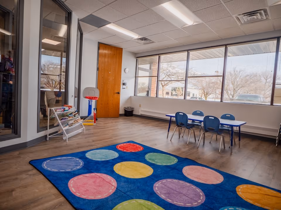 Bright indoor playroom with large windows, colorful rug, and child-sized table and chairs.