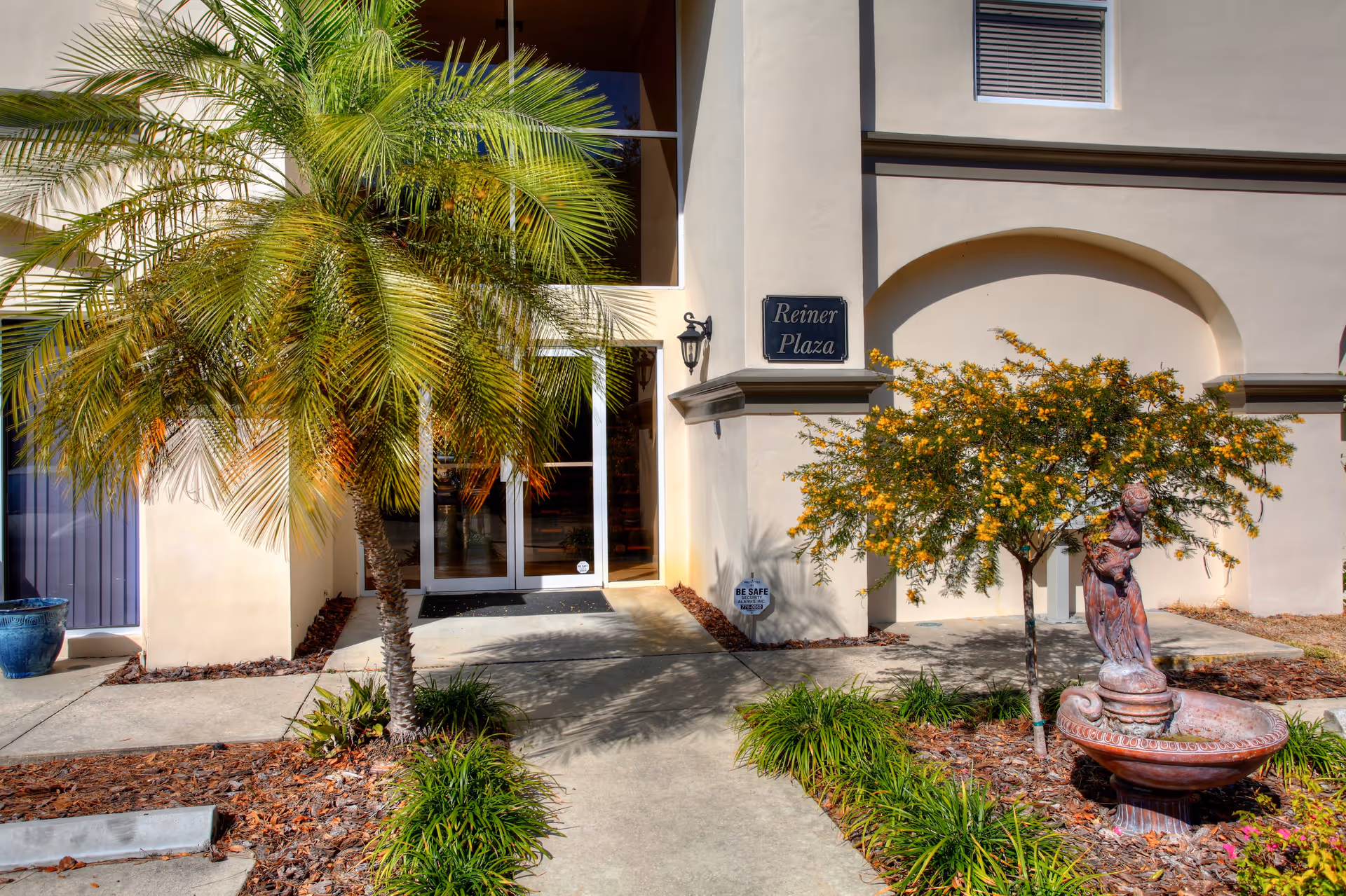 Exterior view of a senior living building entrance with palm trees, statue fountain, and landscaped path.