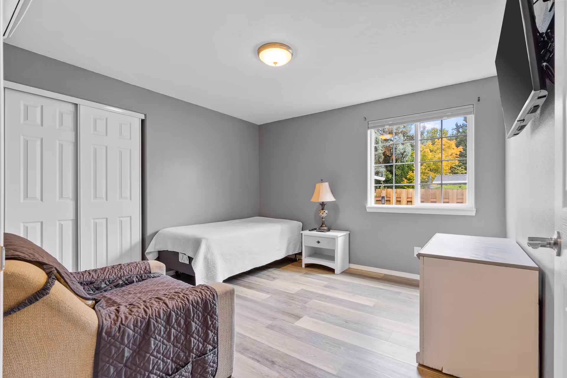 Bedroom in a senior living facility with bed, nightstand, dresser, and window.